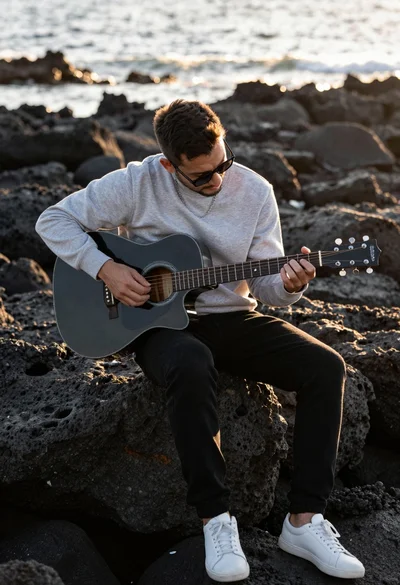 Man playing black acoustic guitar sitting on dark volcanic rocks at sunset
