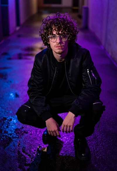 Young man crouching in wet street reflecting purple blue neon lights