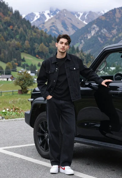 Stylish young man in black denim jacket leaning on suv in mountain