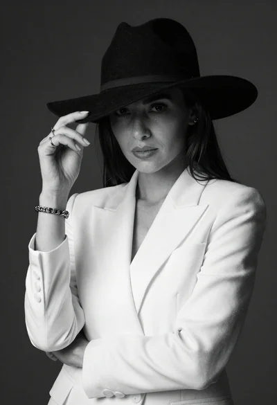 Confident woman in white blazer and wide brimmed hat posing in black