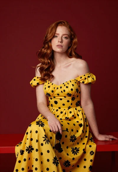 Woman with wavy red hair in yellow floral dress seated on red
