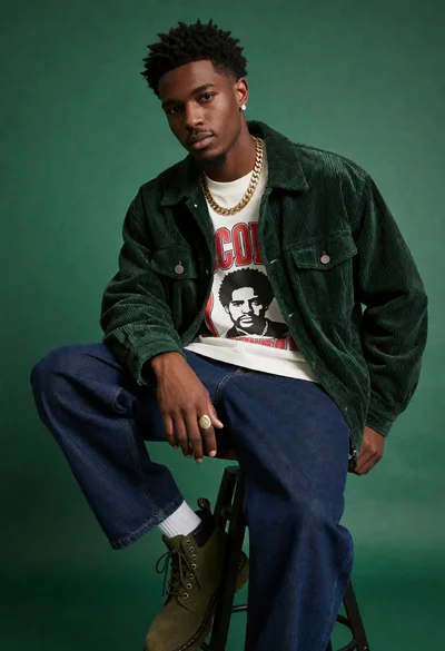 Young black man sitting on stool against solid green backdrop for fashion