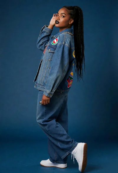 Dark skinned woman in denim jacket posing against solid blue backdrop