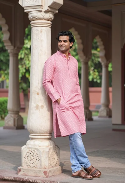 Man in pink kurta leaning against stone pillar outdoors with foliage background