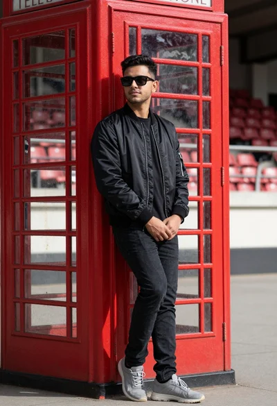 Young man in black jacket leaning against red telephone booth outdoors