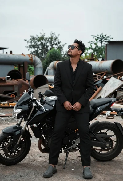 Man in black suit standing beside motorcycle in industrial junkyard setting