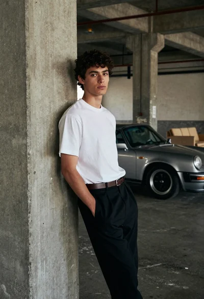 Young man leaning against concrete pillar near classic porsche car