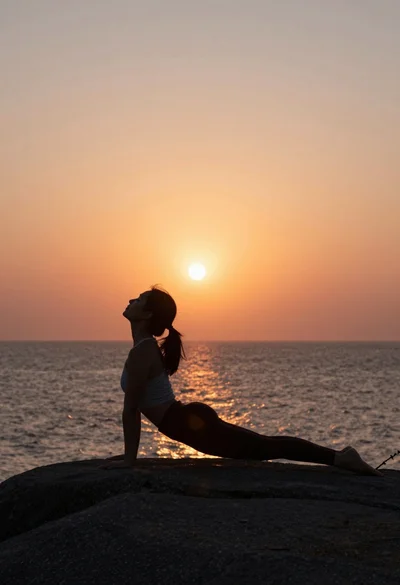 Silhouette of woman doing yoga pose on rocky outcrop at sunset over