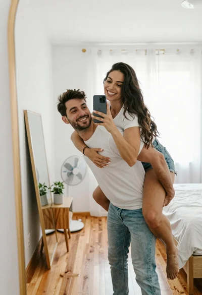 Couple taking mirror selfie in bright bedroom with fan and plant