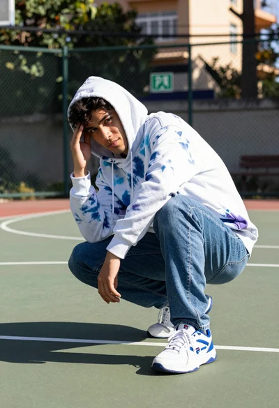 Young man crouching on basketball court wearing tiedye hoodie and jeans