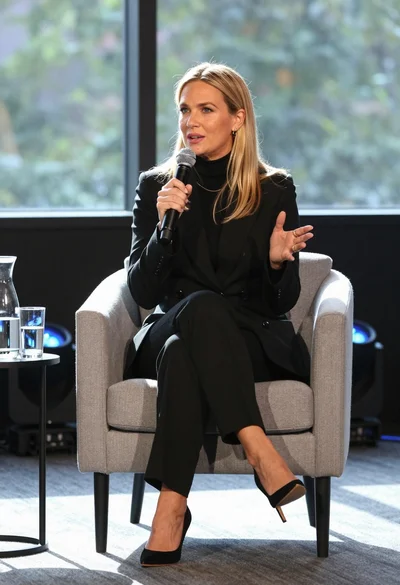 Fair blonde woman in black suit speaking into microphone on stage