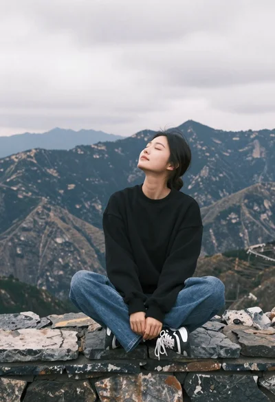 Young woman sitting crosslegged on stone wall with mountains under cloudy sky