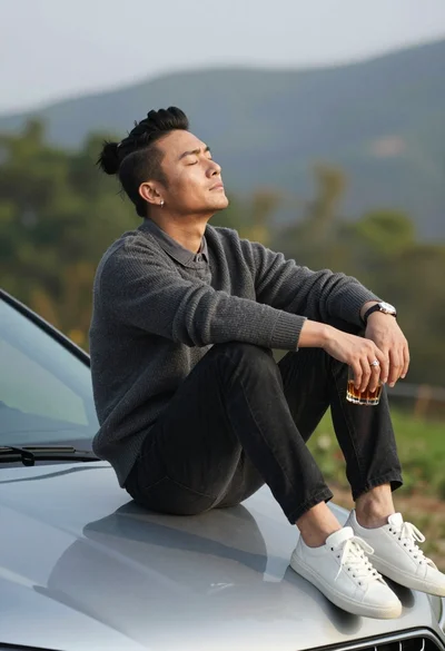 Man sitting on silver car hood outdoors with beverage and hills background