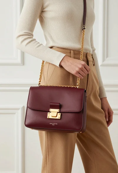 Stylish woman holding burgundy louis vuitton handbag against white paneled wall