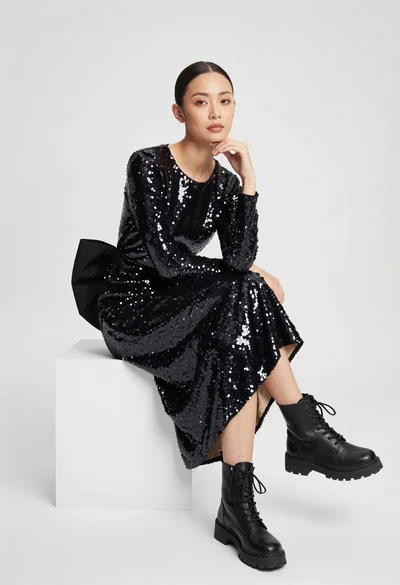 Woman in black sequin dress and combat boots seated in white studio