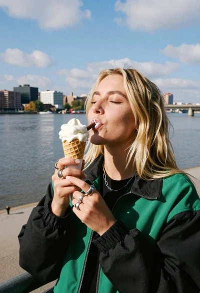 Blonde woman eating ice cream by riverbank with city skyline background