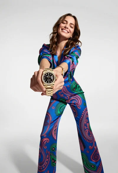 Woman in blue paisley jumpsuit presenting diamond wristwatch against white studio backdrop