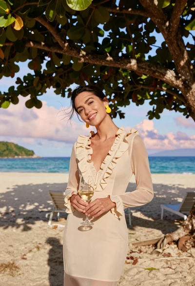 Woman in cream dress holding wine glass on tropical beach under tree