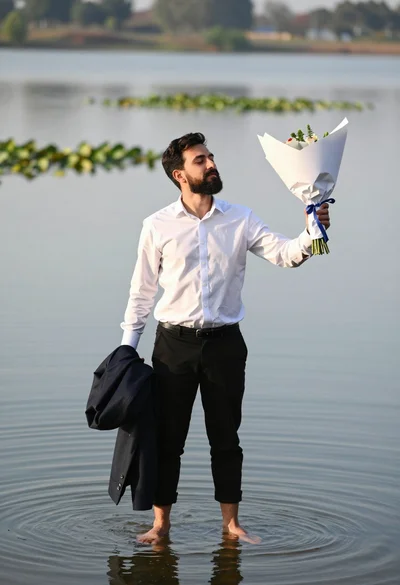 Man standing barefoot in shallow water holding flowers and dark jacket