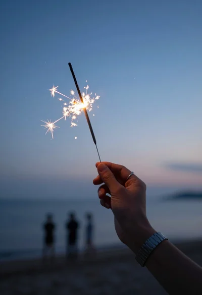 Close up hand holding burning sparkler against a blurred twilight beach sky
