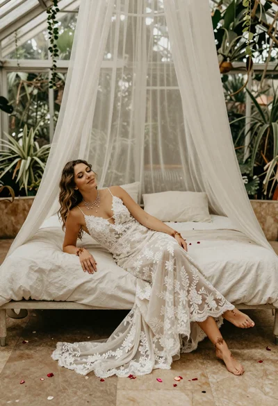 Bride reclining in bohemian lace gown within a sunlit greenhouse atrium