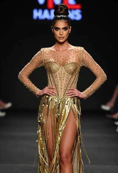 Poised woman in elaborate gold gown on dark runway under spotlight