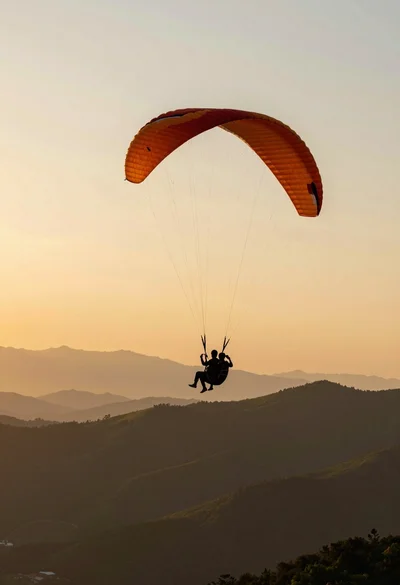 Silhouette of tandem paragliders soaring above green mountain range at sunset