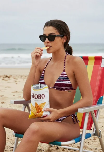 Young woman in patterned bikini eating chips on a sandy beach chair