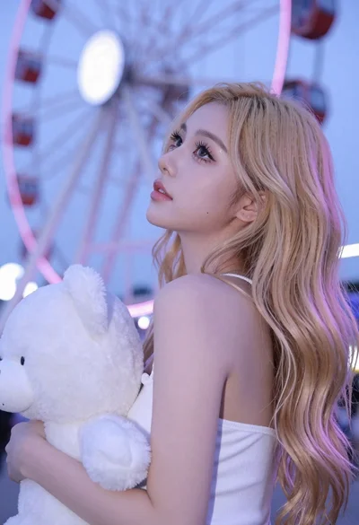 Young blonde woman looking up holding plush toy near ferris wheel lights