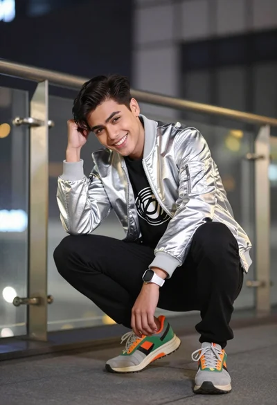 Young man crouching in silver jacket against blurred modern city architecture at