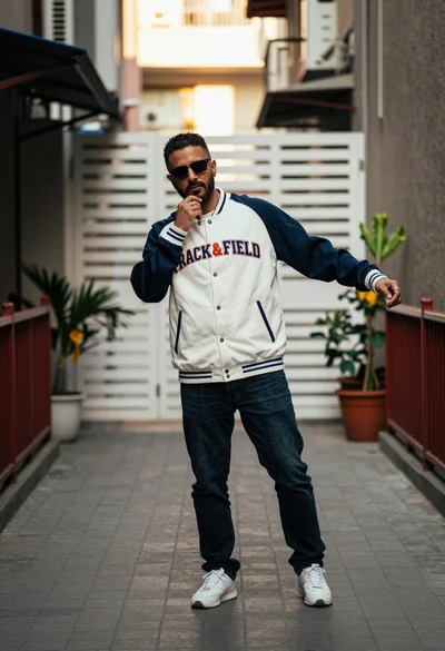Man in varsity jacket posing on paved walkway in sunlit urban alleyway