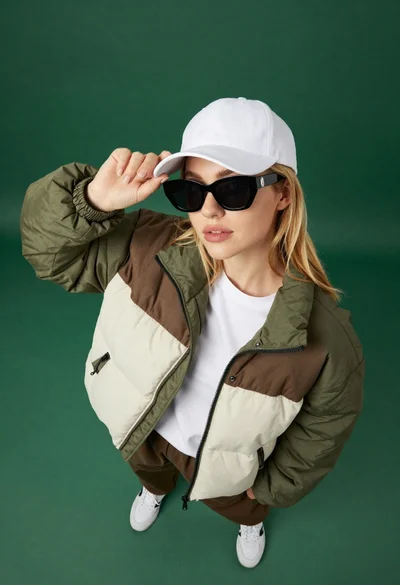 Young woman in colorblocked puffer jacket adjusting white cap from high angle
