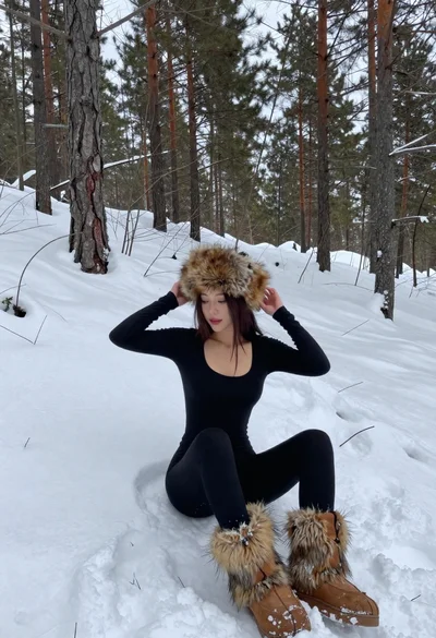 Young woman adjusting fur hat sitting in deep snowy pine forest