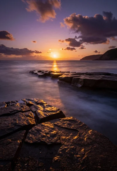Dramatic coastal sunset over calm ocean waters with large foreground rocks