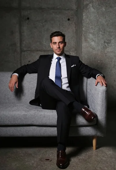 Man in black suit sitting on gray couch against concrete backdrop