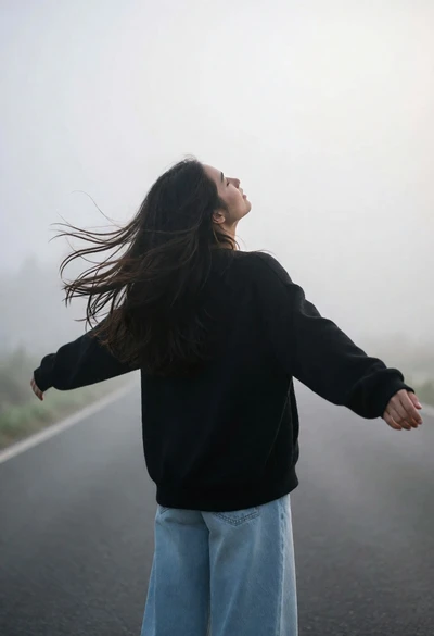 Woman standing on foggy road with outstretched arms looking up into sky