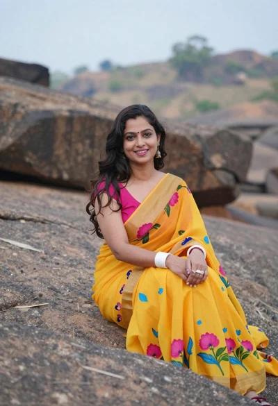 Smiling woman in yellow saree seated on rocks outdoors under overcast sky