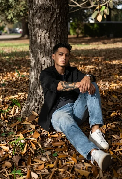 Man with tattoos sitting against a tree trunk in fallen leaf covered