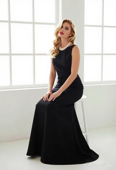 Blonde woman in black gown seated on white stool in bright room