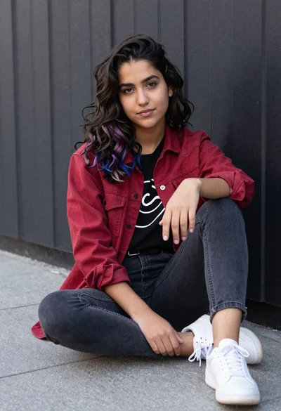 Young woman with dark wavy purple highlighted hair seated on paved surface