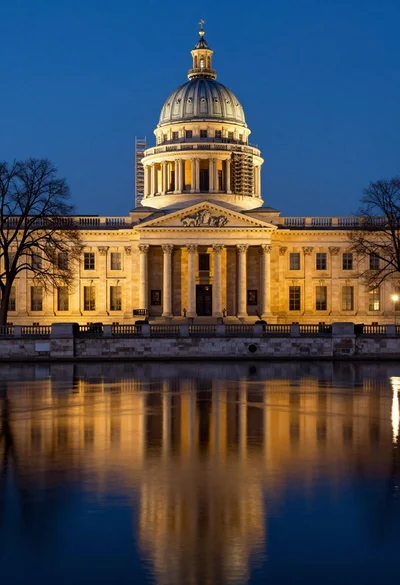 Neoclassical building with dome scaffolding reflecting on calm river water at twilight