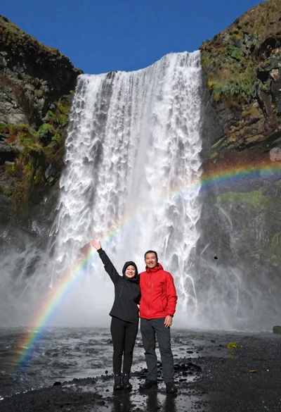 Couple standing before massive waterfall under clear blue sky low angle