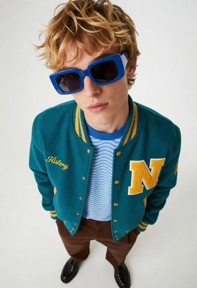 Young man in teal varsity jacket looking up from a white studio