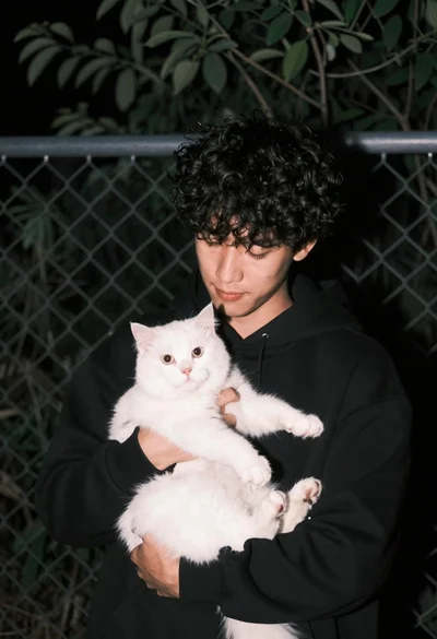 Young man cradling fluffy white cat near chain link fence at night