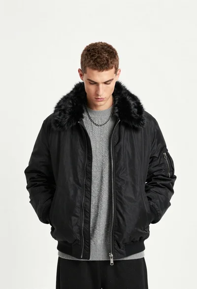 Young man in black bomber jacket and grey sweater standing against white
