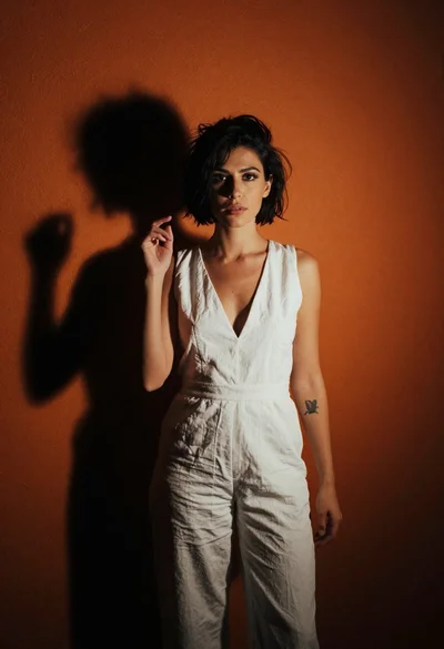 Woman in white jumpsuit against orange stucco wall with dramatic shadows